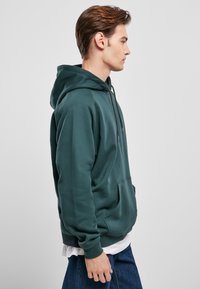 Sudadera verde oscura, hecha de tejido suave, con un bolsillo frontal, capucha con cordón y puños de canalé. Se lleva con una camiseta blanca debajo.