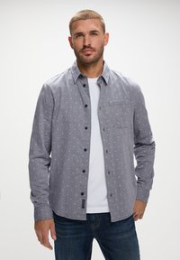 Camisa gris de botones con patrón geométrico blanco, de manga larga, con un bolsillo en el pecho, combinada con una camiseta blanca y jeans azules.