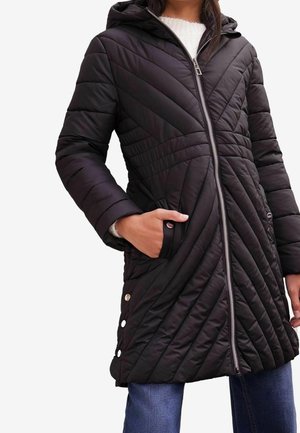 Femme portant un manteau matelassé noir à capuche avec des coutures diagonales, une fermeture éclair à l'avant et des pressions sur le côté, main dans la poche.