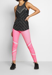 Czarny top na ramiączkach z geometrycznymi szarymi wzorami i logo, w połączeniu z różowymi legginsami z wzorami szachownicy i zygzaków. Czarne sneakersy.