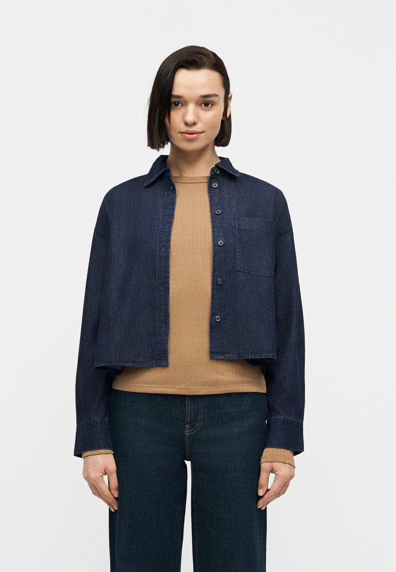 Άτομο με κοντά σκούρα μαλλιά που φοράει ένα κοντό σκουρόχρωμο τζιν μπουφάν πάνω από μια μπεζ ριμπ πλεκτή μπλούζα και σκούρο μπλε τζιν, στέκεται μπροστά σε απλό φόντο.