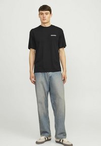 Sort kortærmet t-shirt med hvid logo print på venstre bryst, parret med løse lysvaskede denim jeans og beige sneakers.