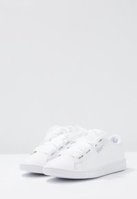 Scarpe sportive bianche con una tomaia sintetica liscia, lacci larghi e suola in gomma testurizzata. Presenta un logo discreto sul lato.