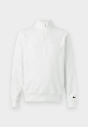 Valge pikkade varrukatega kvart-zip dressipluus püstkraega, soonikkoes mansettide ja alläärega, Lacoste'i logo mansetil ja rinnal asuval plaastril.