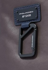 Primer plano de un bolso de cuero oscuro con una etiqueta azul marino cosida que dice "PIQUADRO AP 32436" y un mosquetón geométrico negro adjunto.