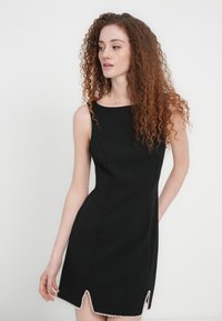 Zwarte nauwsluitende jurk met een ronde halslijn en een textuurrijke afwerking, voorzien van een golvende zoom versierd met kleine witte parels.