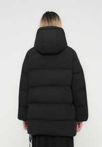 Person mit schulterlangem Haar trägt einen schwarzen gepolsterten Wintermantel mit Kapuze, von hinten vor einem schlichten Hintergrund gezeigt.