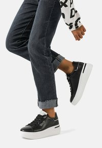 Sneaker in pelle nera con suola piatta bianca, caratterizzati da dettagli texture e design stringato. Indossati con jeans blu scuro con risvolto.