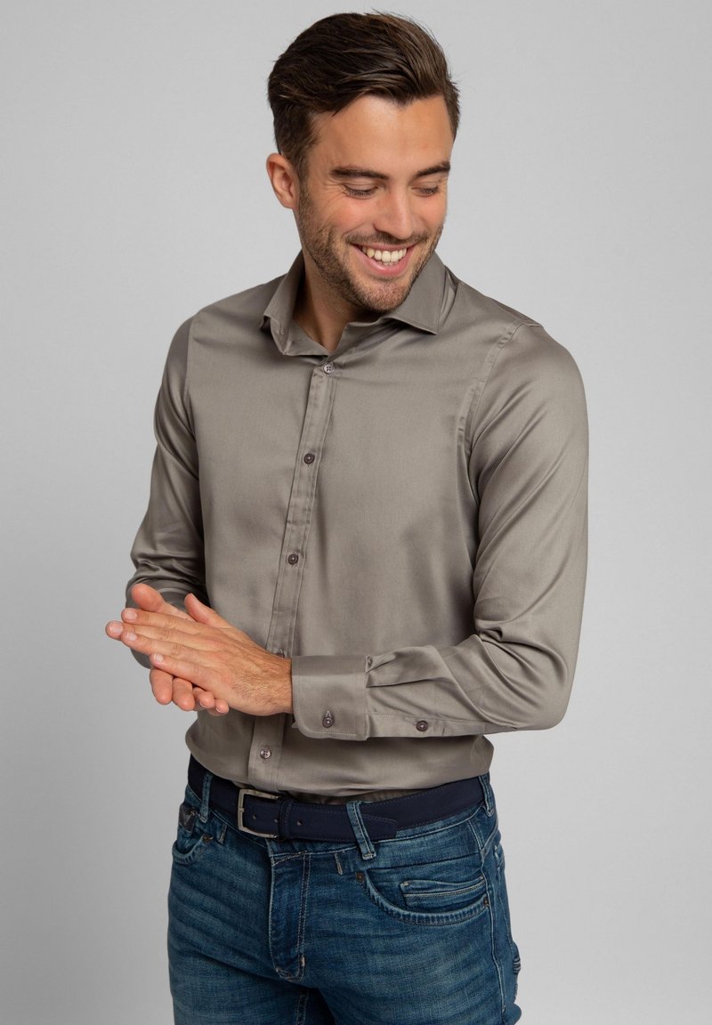 Grijs, langarmig shirt met een gladde textuur, knoopsluiting en donkere knopen. Draagt een blauwe spijkerbroek en een donkere riem.