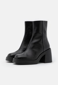 Bottines en cuir noir avec des talons épais et une finition lisse, positionnées côte à côte sur un fond blanc.