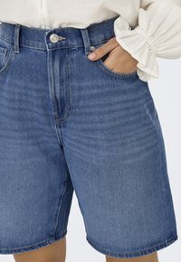 Denim kratke hlače imajo visok pas z gumbom. Tkanina je srednje modre barve z rahlim bledenjem, vidna sta dva stranska žepa.