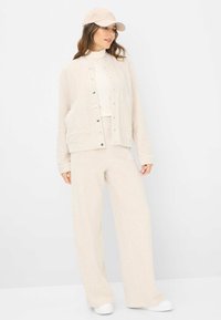 Hellbeige Jacke mit Druckknöpfen und Vordertaschen, kombiniert mit passenden weiten Hosen und einer passenden Mütze. Glatte Stoffstruktur.