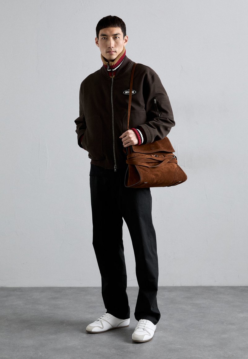 Brun zip-up-jacka med ribbad krage; svarta byxor; vita sneakers. Brun mockaväska med lager av remmar och sömnadsdetaljer.