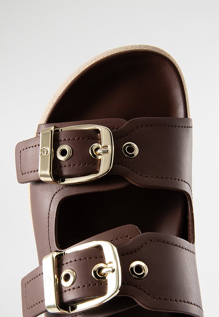 Braune Leder-Sandale mit zwei verstellbaren Riemen, die goldfarbene Schnallen aufweisen. Flache Sohle mit einem strukturierten, hellen Boden.