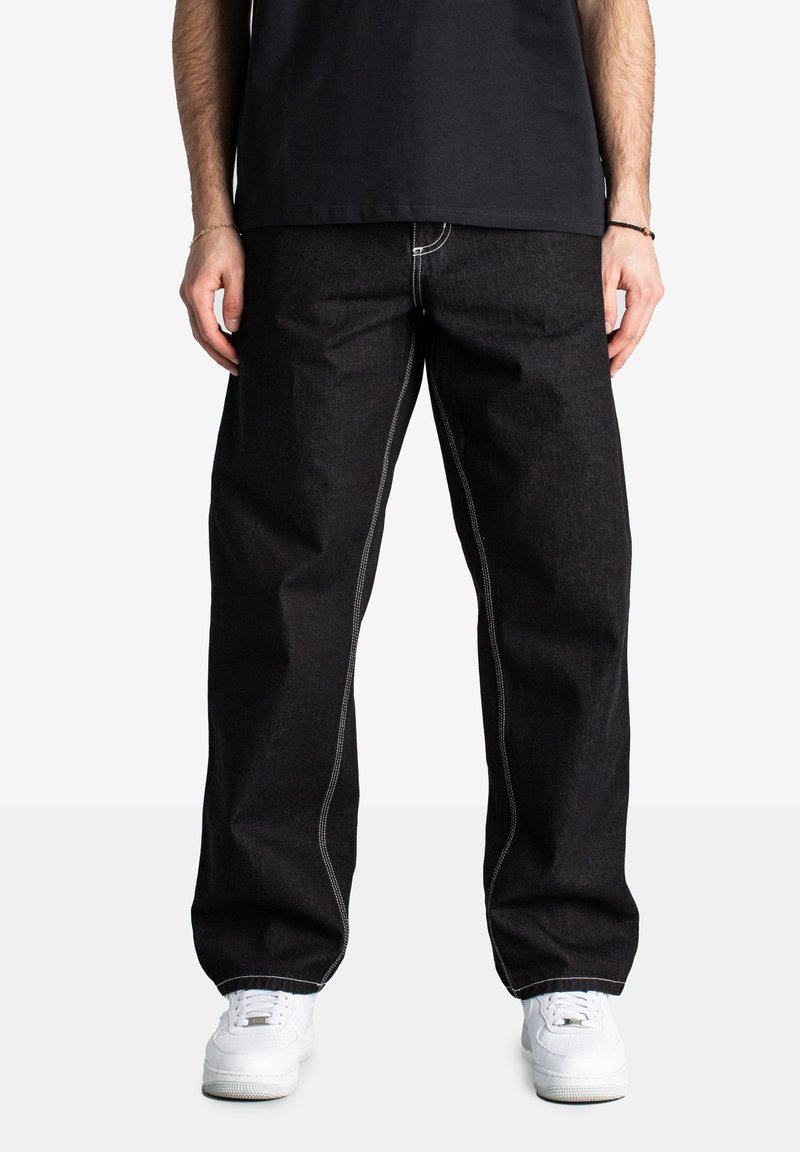 Jeans in denim nero con gambali larghi, caratterizzati da cuciture bianche in contrasto lungo le cuciture. Indossati con sneakers bianche e una maglietta nera.