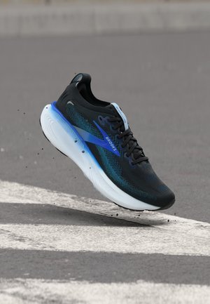 Zapatilla de correr Brooks negra y azul en el aire sobre una superficie de asfalto gris con rayas blancas y partículas de suciedad alrededor.