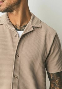 Puff-sleeved beige collared shirt with a textured ribbed fabric, featuring two buttons and a plain white undershirt visible beneath.