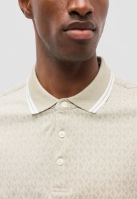 Gros plan d’un homme portant un polo beige avec un motif de logo MK et un col rayé blanc, montrant les boutons et le bas du visage.