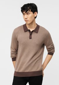 Νέος άνδρας με κοντά μαύρα μαλλιά που φοράει ανοιχτή καφέ μπλούζα πόλο με μακριά μανίκια και μαύρο παντελόνι, στέκεται μπροστά σε λευκό φόντο.