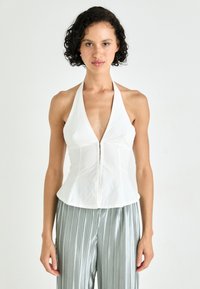 Top blanco sin mangas con escote en V profundo y cierre frontal con cremallera, que presenta un cuerpo ajustado y un dobladillo acampanado. Textura de tela suave y lisa.