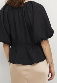Black, textured blouse with puffed sleeves, gathered waist, and flowing hem. Designed for a relaxed fit, featuring a simple, elegant silhouette.