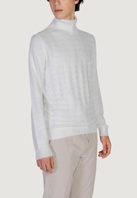 Bijela turtleneck majica s teksturiranim dijamantnim uzorkom. Mekani materijal, uski kroj, rebrasti rukavi i donji rub. Uparena s svijetlo bež hlačama.