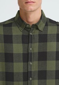 Camicia a quadri verdi e neri con colletto a bottoni, caratterizzata da un tessuto testurizzato e sei bottoni lungo la patta centrale.