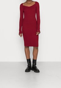 Robe en maille rouge côtelée à manches longues, encolure dégagée et longueur midi, associée à des bottines noires. Motif texturé visible.