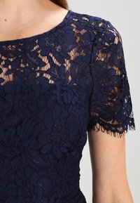 Abito in pizzo floreale blu navy con maniche corte, corpino semi-trasparente e intricate fantasie floreali su tessuto trasparente.