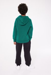 Felpa verde con cappuccio, maniche lunghe e orlo inferiore. Abbinata a pantaloni da tuta neri e sneakers bianche. Tessuto morbido, vestibilità rilassata.