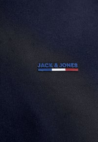 Sötétkék szövet, rajta "JACK & JONES" felirat egy vízszintes csík felett, amely kék, fehér és piros színekből áll.