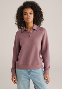 Pulover majica u boji maline s ovratnikom, kopčanjem na gumbe i raglan rukavima; izrađena od mekanog, teksturiranog tkanja.