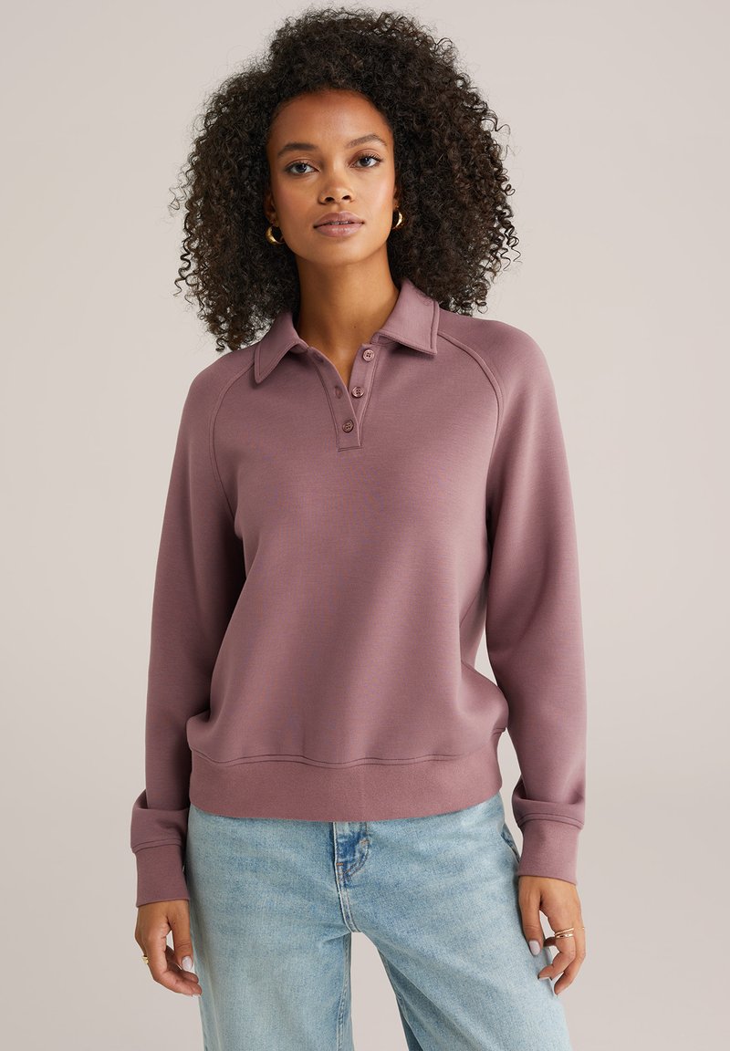 Pulover majica u boji maline s ovratnikom, kopčanjem na gumbe i raglan rukavima; izrađena od mekanog, teksturiranog tkanja.