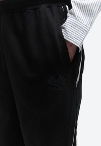 Άτομο που φοράει μαύρο παντελόνι φόρμας με το λογότυπο "ravela" στο μηρό, χέρι στην τσέπη, και λευκό πουκάμισο με μπλε ρίγες ορατές στο μανίκι.