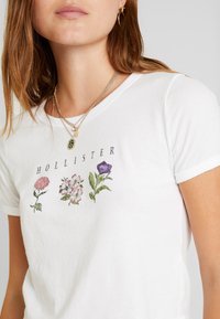 White cotton t-shirt with short rolled sleeves, featuring embroidered floral designs in pink, purple, and green, and the text "HOLLISTER."