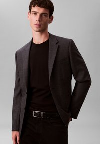 Blazer gris foncé ajusté avec une fermeture à deux boutons, une légère texture et une poche poitrine unique, porté sur un T-shirt noir et un jean foncé.