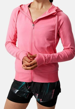 Femme portant un sweat à capuche zippé rose vif et un short de sport noir à motifs, debout les mains jointes devant.