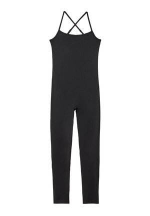 Vrouw die een strak, mouwloos zwart jumpsuit met dunne bandjes aanpast tegen een effen lichte achtergrond.