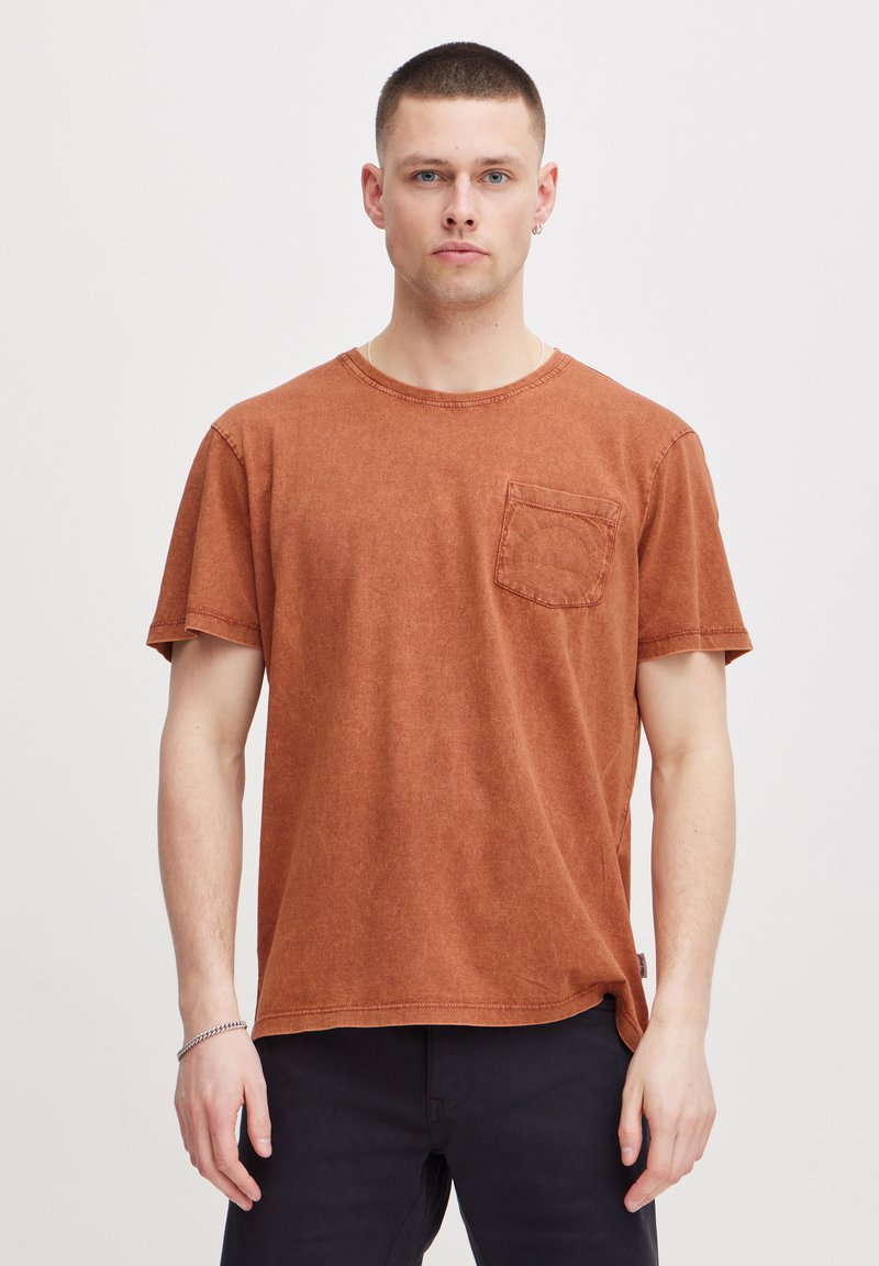 Camiseta de algodón color óxido con mangas cortas, cuello redondo y bolsillo frontal. Presenta un corte holgado y una textura sutil.
