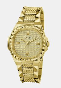 Reloj Guess en tono dorado con una pulsera texturizada y tachonada. La esfera presenta cristales brillantes y marcadores horarios en relieve, con forma octagonal.