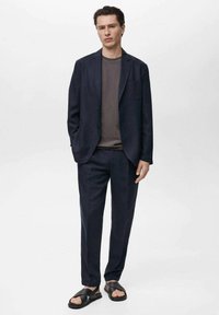 Costum din blazer și pantaloni marine, cu un top gri. Țesătură texturată, croială ajustată și sandale negre cu talpă plată și bretele încrucișate.