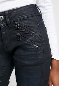 Svarta denimbyxor med flera diagonala dragkedjor framtill, en slät textur och en åtsittande design.