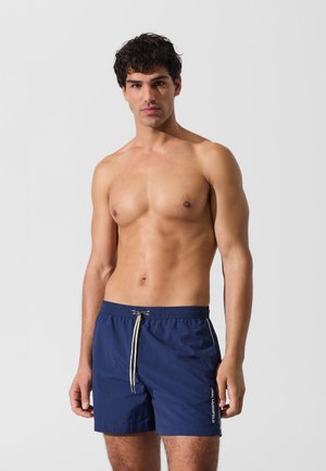 Tmavomodré plavecké šortky s elastickým pásom a šnúrkou. Majú biely detail loga na boku a rovný strih. Hladká textúra.
