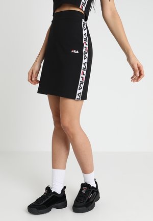 Vrouw draagt zwarte Fila-rok met zijlogo strepen, zwarte Fila-sneakers en witte sokken, staand tegen een effen achtergrond.
