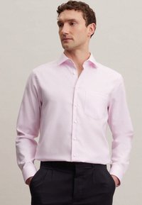 Camicia rosa chiaro a maniche lunghe con colletto a punta, tasca frontale e bottoni bianchi. Indossata con pantaloni scuri, che mettono in mostra un design aderente.