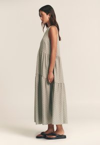 Maxi obleka iz mehkega sivega materiala s črnimi pikami, z večslojno zasnovo in brezrokavnikom. Kombinirano s črnimi sandalami.