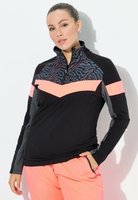Donna in abbigliamento sportivo nero e rosa neon con stampa animale sul petto, in piedi contro un muro bianco con una mano in tasca.