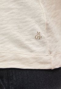 Μπεζ ύφασμα με λεπτή υφή πλέξης και μικρά γράμματα "M O P" κοντά στη ραμμένη άκρη πάνω από σκούρο τζιν ύφασμα.