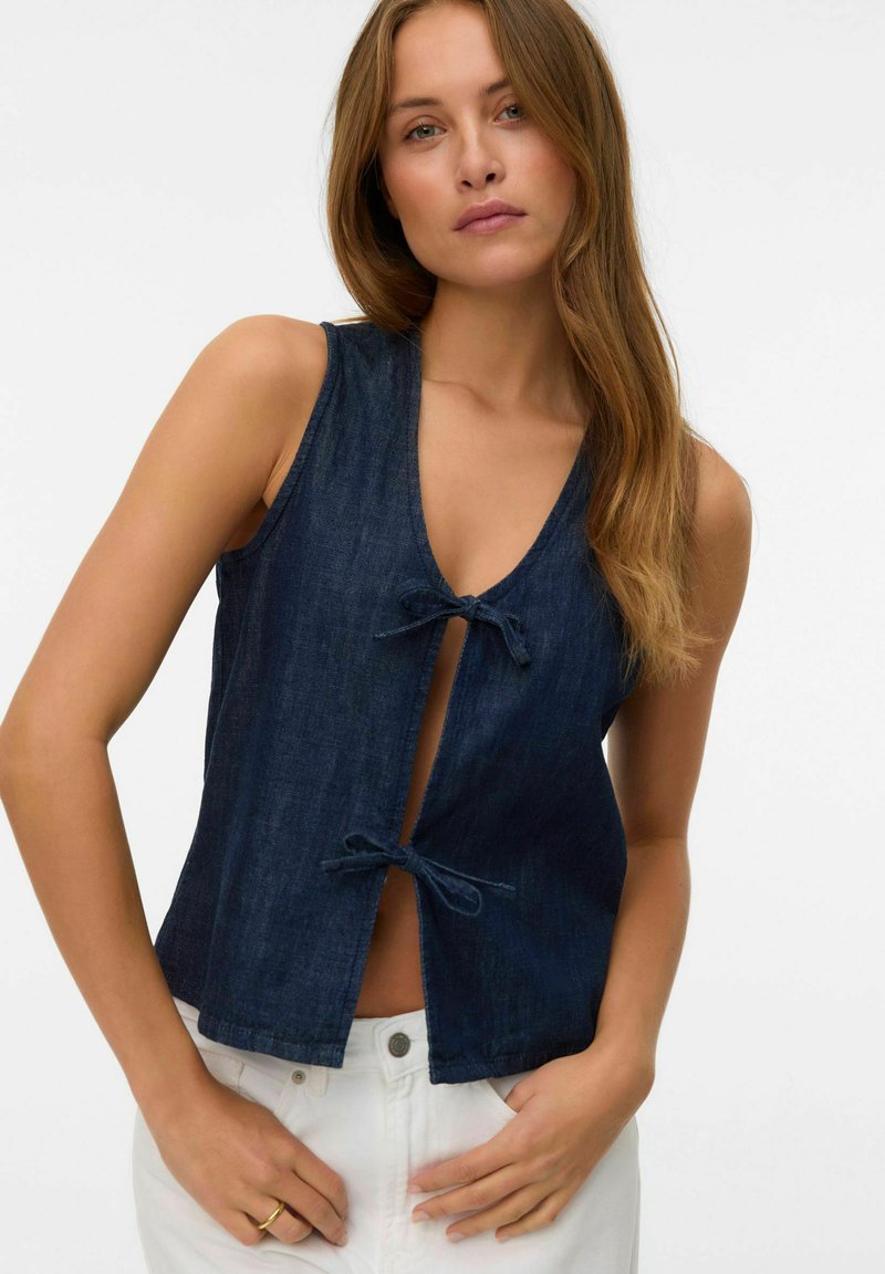 Mujer con cabello largo que lleva un chaleco sin mangas de mezclilla oscura abrochado con dos lazos delanteros y pantalones blancos, posando con las manos cerca de los bolsillos.