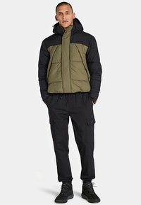 Kurtka puchowa o zielonym korpusie i czarnych ramionach, z pełnym zamkiem błyskawicznym i kapturem. Połączona z czarnymi spodniami typu cargo i ciemnym obuwiem.